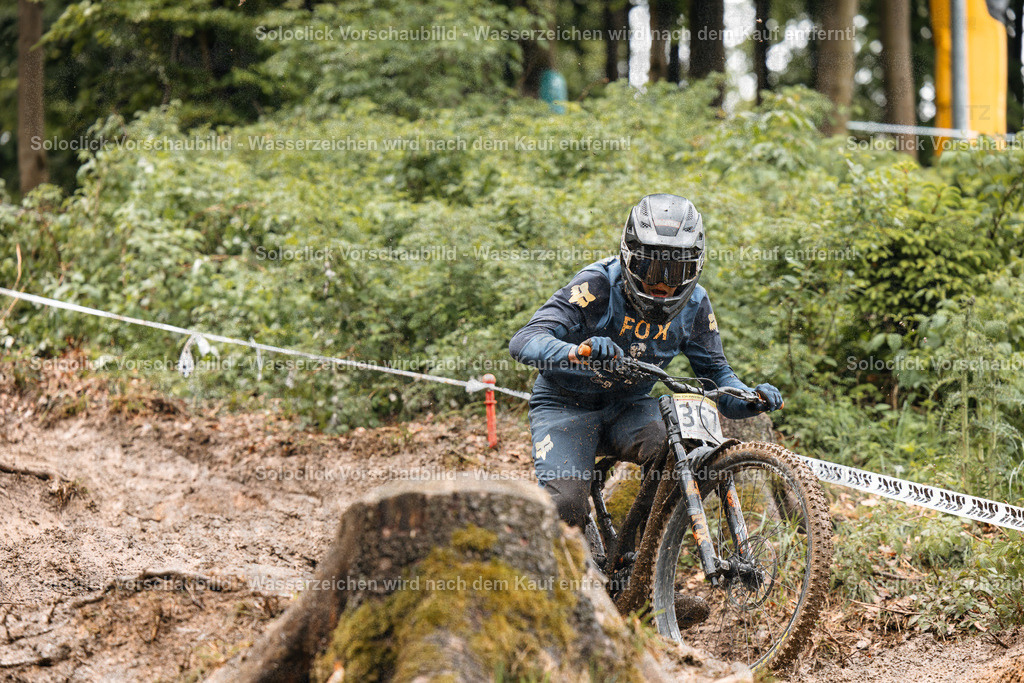 Mountainbike-Abenteuer im Wald | Ein Mountainbiker in voller Ausrüstung rast einen staubigen, kurvigen Trail hinunter. Er trägt einen schwarzen Helm mit orangefarbenen Akzenten und eine dunkle Schutzbrille. Sein Trikot und seine Hose sind ebenfalls schwarz und tragen das Logo von Fox Racing. Er trägt eine Nummer 30 auf dem Rücken. Der Trail schlängelt sich durch einen dichten Wald, der mit dichtem Unterholz und Bäumen bedeckt ist. Der Trail ist mit roten und weißen Markierungen versehen. Der Himmel ist bewölkt. - Realisiert mit Pictrs.com
