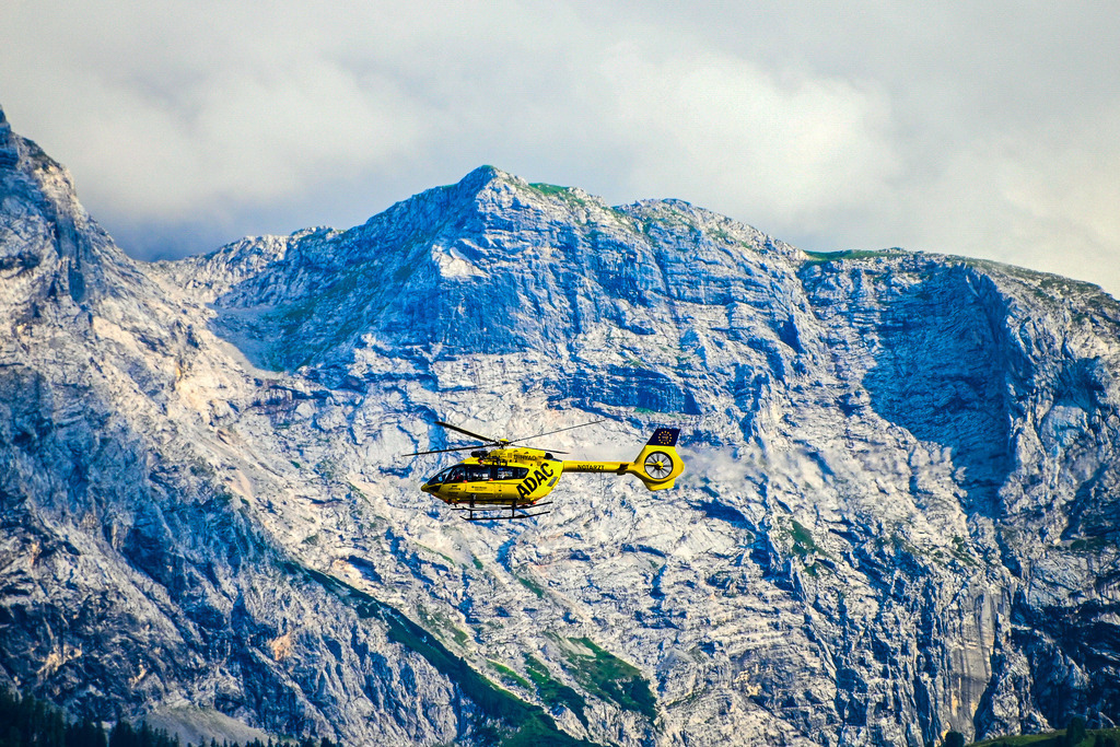 Engel in Gelb: Hoffnung über den Wolken | Ein Bild, das den Atem anhalten lässt und tief bewegt: Vor der gigantischen, unbarmherzig schroffen Felswand des Bergmassivs zieht ein gelber ADAC-Rettungshubschrauber seine Bahnen. Winzig wirkt die Maschine vor der monumentalen Kulisse aus Stein und den heraufziehenden, dichten Wolken – und ist doch ein mächtiges Symbol für Rettung, Hoffnung und menschliche Hilfsbereitschaft in extremen Momenten. Die Aufnahme fängt die dramatische Spannung zwischen der Urgewalt der Alpen und dem unermüdlichen Einsatz der Luftretter perfekt ein. Ein Bild voller Emotion, Ehrfurcht und Dankbarkeit. - Realisiert mit Pictrs.com