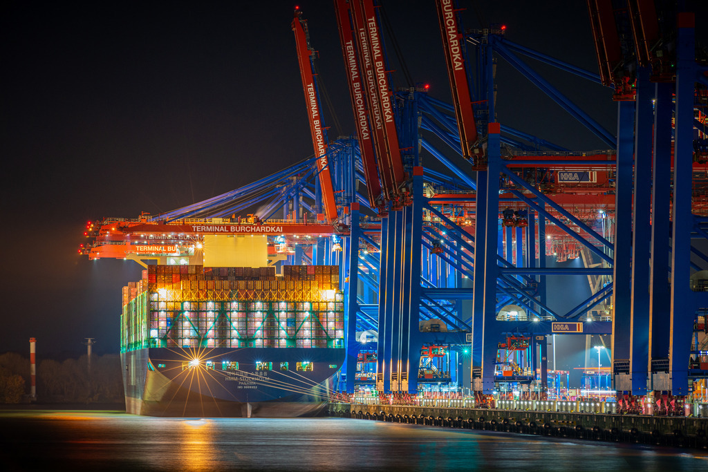 Nachts am Burchardkai Nr.2 | Das Bild zeigt eine nächtliche Szenerie im Hamburger Hafen am Burchardkai, wo mehrere riesige Containerschiffe angedockt sind. Die Schiffe erheben sich majestätisch in die Dunkelheit und werden von einer Flut von Lichtern beleuchtet, die vom Hafen und von den Schiffen selbst ausgehen. Das Bild fängt die Hektik und den Puls des Hafens in der Nacht ein und vermittelt einen Eindruck von der enormen Größe und dem Einfluss dieses wichtigen Handelszentrums.