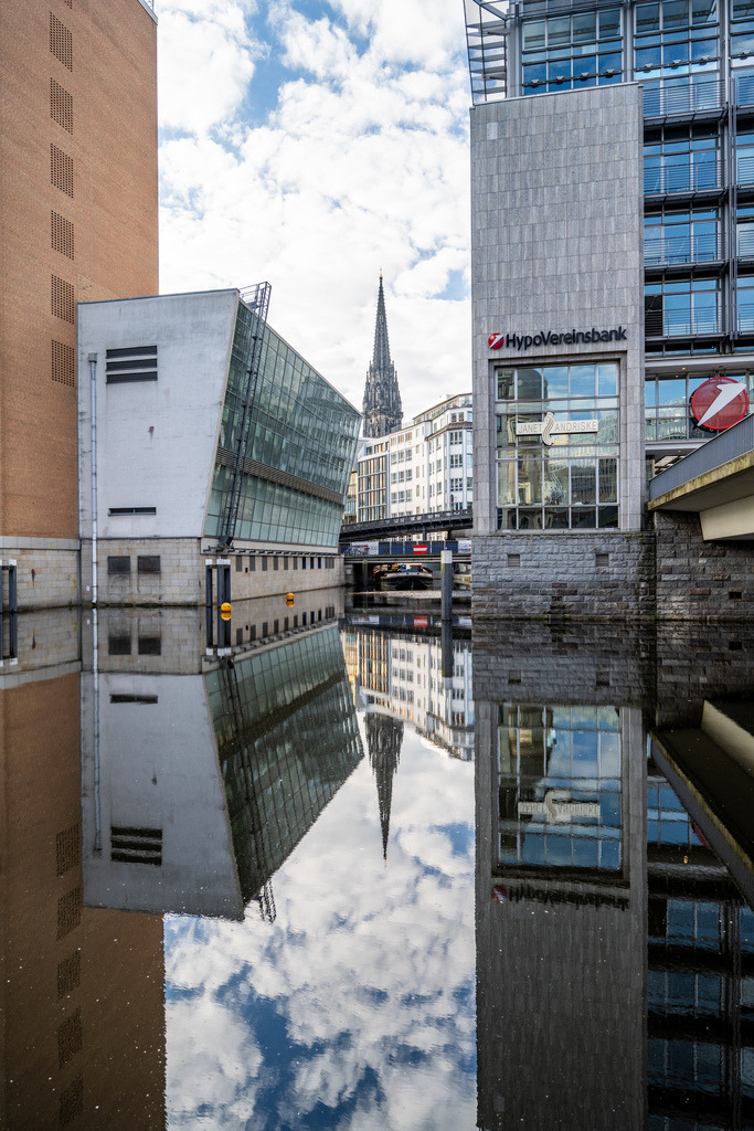 Hamburger Architektur zwischen Himmel und Spiegelung | Eine moderne Hamburger Gebäudefassade spiegelt sich gemeinsam mit dem Himmel in der ruhigen Wasserfläche. Das Hochformat betont die vertikalen Linien und die grafische Wirkung der Architektur. - Realisiert mit Pictrs.com