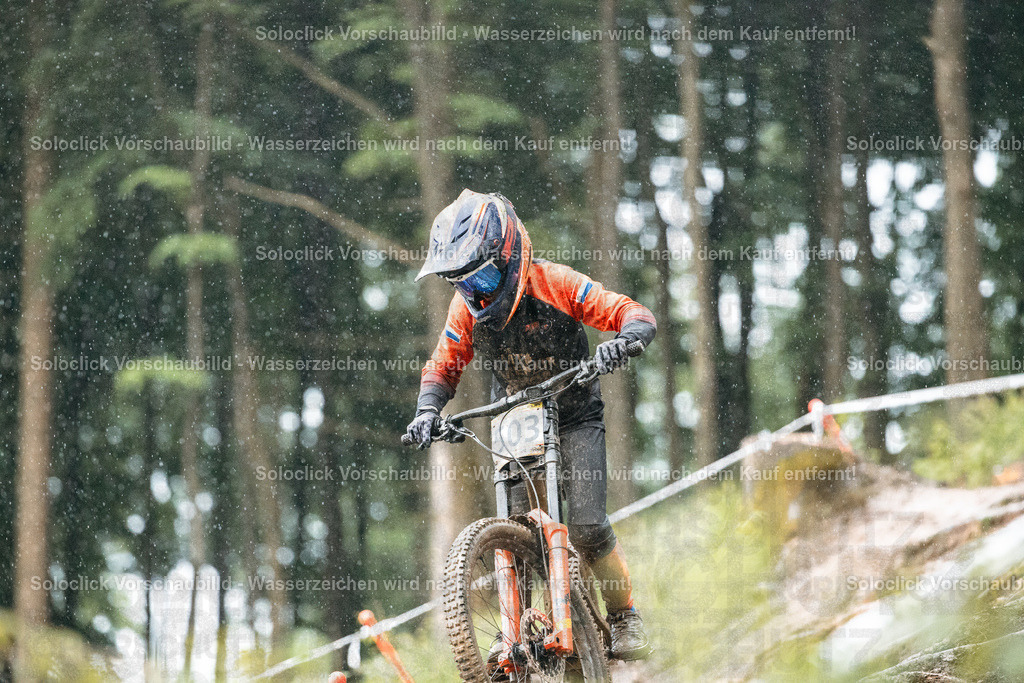 Mountainbiker im Rennen | Ein Mountainbiker in voller Ausrüstung rast einen steilen, verschlammten Trail hinunter. Der Fahrer trägt einen orangefarbenen Helm und eine Jacke, sowie Handschuhe und eine Schutzbrille. Er trägt die Nummer 03 auf seinem Rücken. Der Trail ist mit Markierungskegeln versehen, die den Weg weisen. Der Hintergrund besteht aus dichtem Grünwerk, hauptsächlich Bäume. - Realisiert mit Pictrs.com