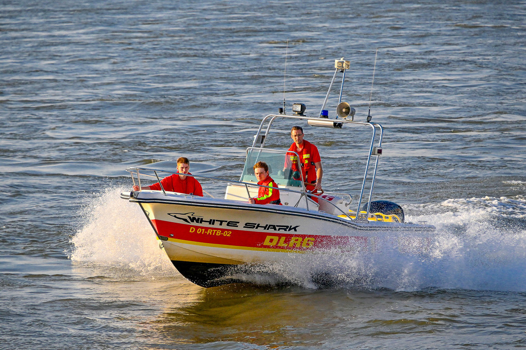 Einsatz auf den Wellen: Die DLRG in Aktion | Das Bild zeigt ein Rettungsboot der DLRG mit dem Namen „White Shark“ (Kennung D 01-RTB-02), das mit hoher Geschwindigkeit über den Rhein bei Düsseldorf fährt. Am Bug und Steuerstand sind drei Einsatzkräfte in ihrer typischen rot-gelben Schutzkleidung zu sehen. Das Boot schneidet dynamisch durch das Wasser, wodurch eine markante Gischt- und Heckwelle entsteht. Das warme Sonnenlicht des späten Nachmittags beleuchtet die Szene und lässt das Wasser in tiefen Blautönen schimmern. - Realisiert mit Pictrs.com