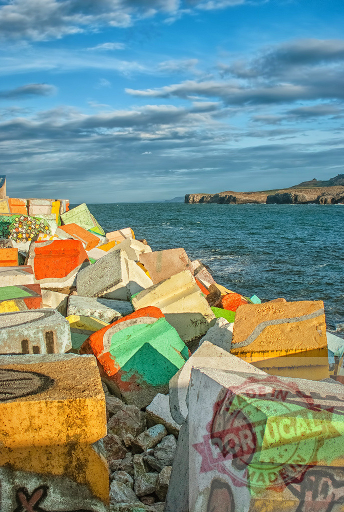 Colorful concrete blocks forming breakwater protecting coastline from erosion | Brightly painted concrete blocks creating breakwater along rocky shore, protecting against coastal erosion under cloudy sky - Realized with Pictrs.com