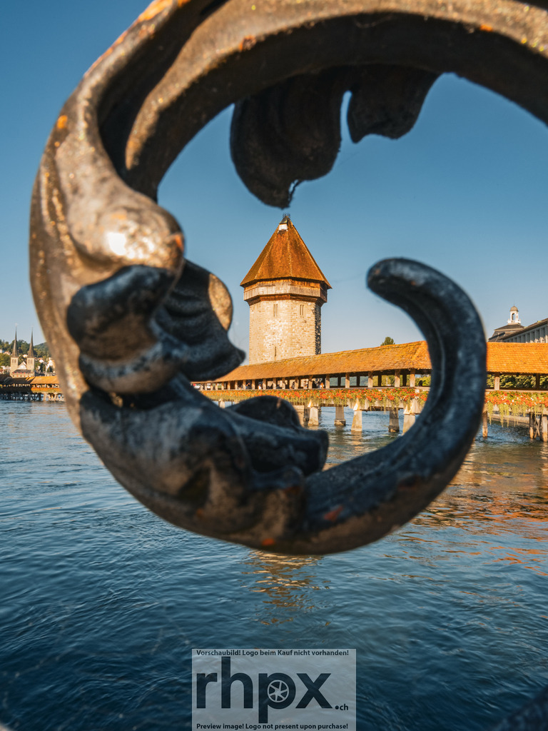 Kapellbrücke Luzern Fokus | Geschichte. Rahmen. Klarheit.Der Blick durch das schmiedeeiserne Ornament lenkt den Fokus direkt auf den historischen Wasserturm und die hölzerne Kapellbrücke. Die kräftigen Farben des Wassers und der Architektur fangen die zeitlose Atmosphäre der Stadt Luzern perfekt ein. Ein Spiel aus Form und Tradition.Wähle unter "Produktauswahl" dein Wunschformat: Vom klassischen Wandbild über Puzzle &amp; Tassen bis zum digitalen Download (z.B. als Handy-Hintergrund). - Realisiert mit Pictrs.com