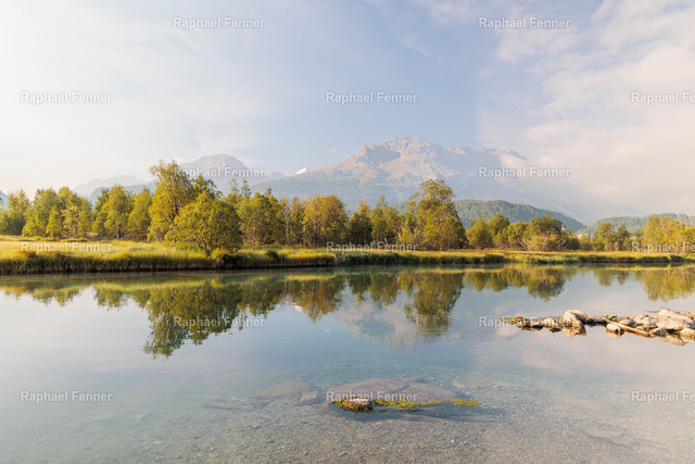 Spiegelung am Inn bei Samedan | Ein ruhiger Morgen am Inn bei Samedan im Oberengadin. Das warme Sonnenlicht taucht die Landschaft in sanfte Farben, während sich die Bergkulisse und die Bäume nahezu perfekt im klaren Wasser spiegeln. Die friedliche Atmosphäre macht dieses Motiv zu einem zeitlosen Blickfang für Wohn- und Arbeitsräume.