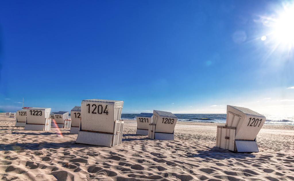 Kultige Strandkörbe an Strand von Westerland in Sylt--Meer | Kultige Strandkörbe: Im Vordergrund stehen mehrere weiße Strandkörbe mit markanten Nummern wie 1204, 1201 und 1203. Die typisch eckige Form der Körbe deutet klar auf das klassische Nordseedesign hin.Landschaft: Der feine, helle Sandstrand zieht sich bis an die Nordsee. Im Hintergrund sind sanfte Meereswellen unter einem strahlend blauen Himmel zu sehen.Atmosphäre: Die strahlende Sonne oben rechts erzeugt eine warme Urlaubsstimmung und wirft sanfte Schatten auf die wellige Sandstruktur. - Realisiert mit Pictrs.com
