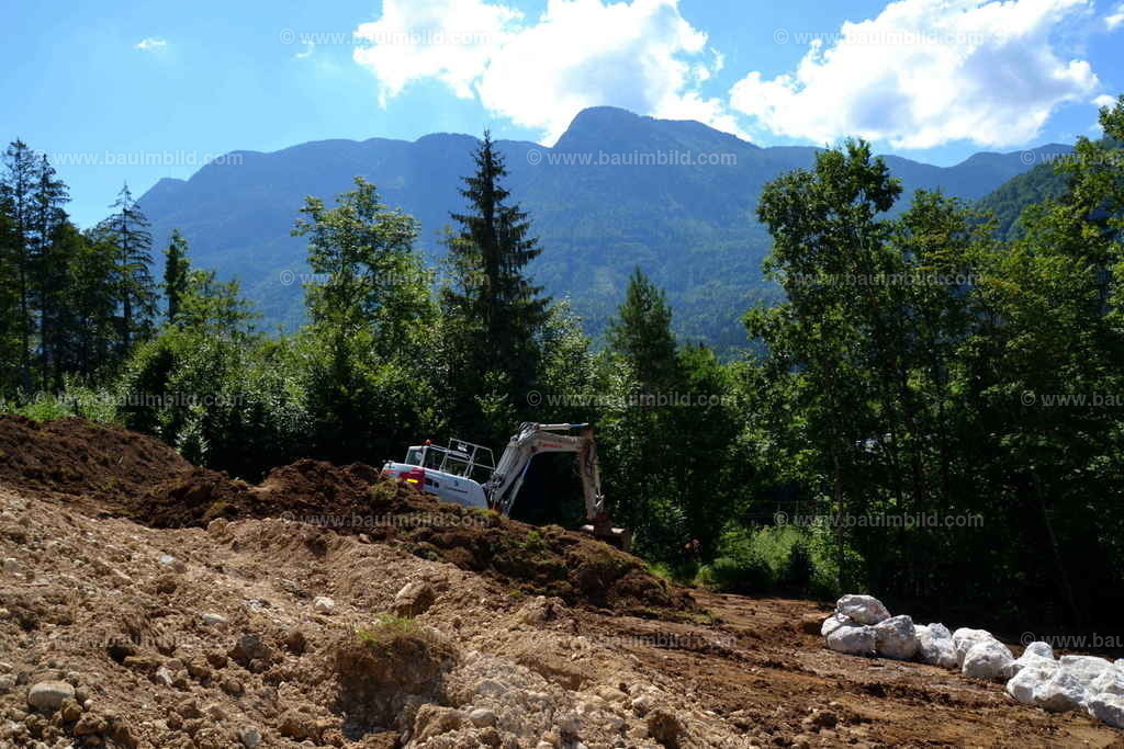 Erdarbeiten mit Bagger | Mittelgroßer Raupenbagger auf Baugrund in Hanglage, gelagerte Kalksteine für Steinwurf, Hintergrund Berge, blauer Himmel mit Wolken