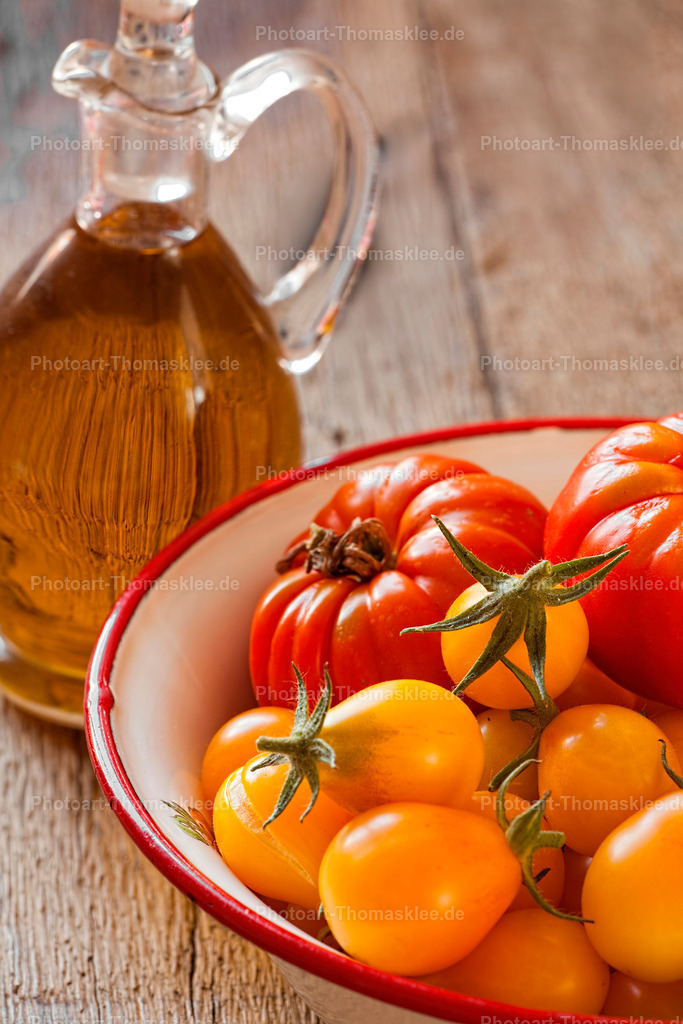 Bunte Biotomaten und Olivenöl auf rustikalem Holz | Eine weiße Emailleschale mit rotem Rand präsentiert eine verlockende Auswahl frischer Biotomaten in verschiedenen Größen und Farben. Leuchtend rote Fleischtomaten harmonieren perfekt mit goldgelben Cherrytomaten, deren grüne Stiele noch die Frische unterstreichen. Eine elegante Glaskaraffe mit goldenem Olivenöl steht im Hintergrund auf der rustikalen Holzoberfläche. Diese mediterrane Komposition verkörpert die Essenz der gesunden Küche und lädt zum Kochen ein. - Realisiert mit Pictrs.com
