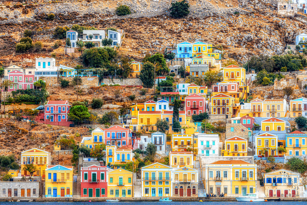 Die charakteristischen, bunten neoklassischen Häuser im malerischen Hafenort der griechischen Insel Symi | Das Bild zeigt die charakteristischen, bunten neoklassischen Häuser im malerischen Hafenort der griechischen Insel Symi (Dodekanes).Architektur: Die Häuser sind in Pastellfarben gehalten und terrassenförmig an die steilen Hänge rund um den Naturhafen (Gialos) gebaut.Lage: Die Insel liegt in der südöstlichen Ägäis, nur wenige Kilometer nördlich von Rhodos.Sehenswürdigkeiten: Neben dem Haupthafen ist das im Süden gelegene, historische Kloster des Erzengels Michael Panormitis ein beliebtes Ausflugsziel - Realisiert mit Pictrs.com