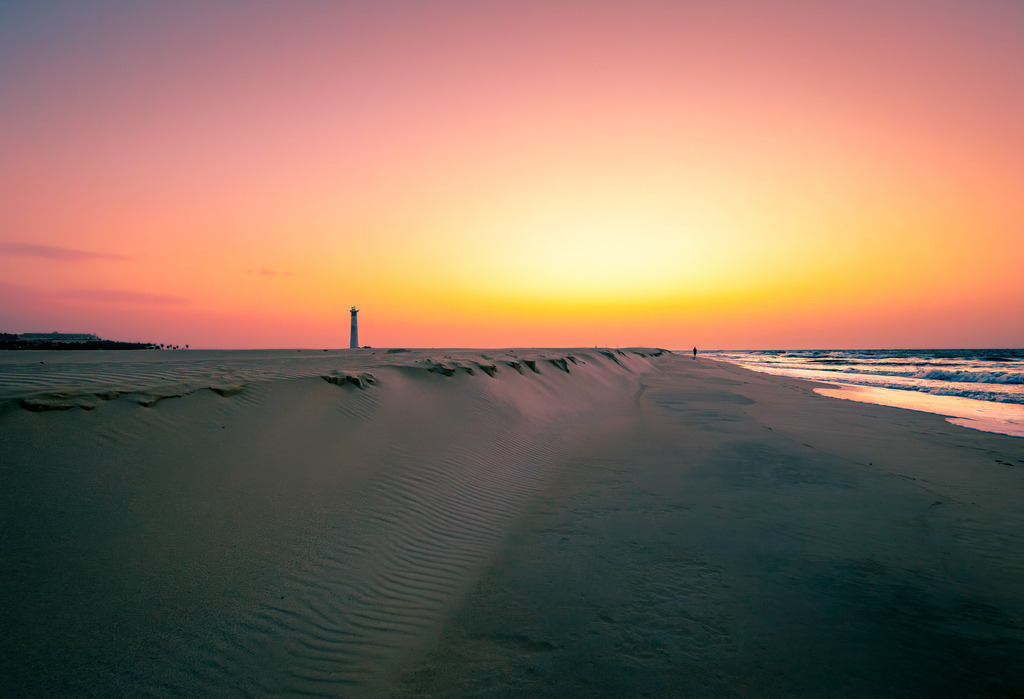 Sonnenaufgang am Strand Jandia--Leuchtturm---Fuerteventura | Das Bild zeigt einen ruhigen Strand on Jandia in Fuerteventura bei Sonnenaufgang.Motiv: Ein Sandstrand erstreckt sich neben dem Meer, während die Sonne am Horizont untergeht und den Himmel in warme Orange- und Rottöne taucht.Details: In der Ferne ist ein Leuchtturm erkennbar, und auf dem Wasser sind sanfte Wellen zu sehen.Atmosphäre: Die Szene vermittelt ein Gefühl von Ruhe und Entspannung. - Realisiert mit Pictrs.com
