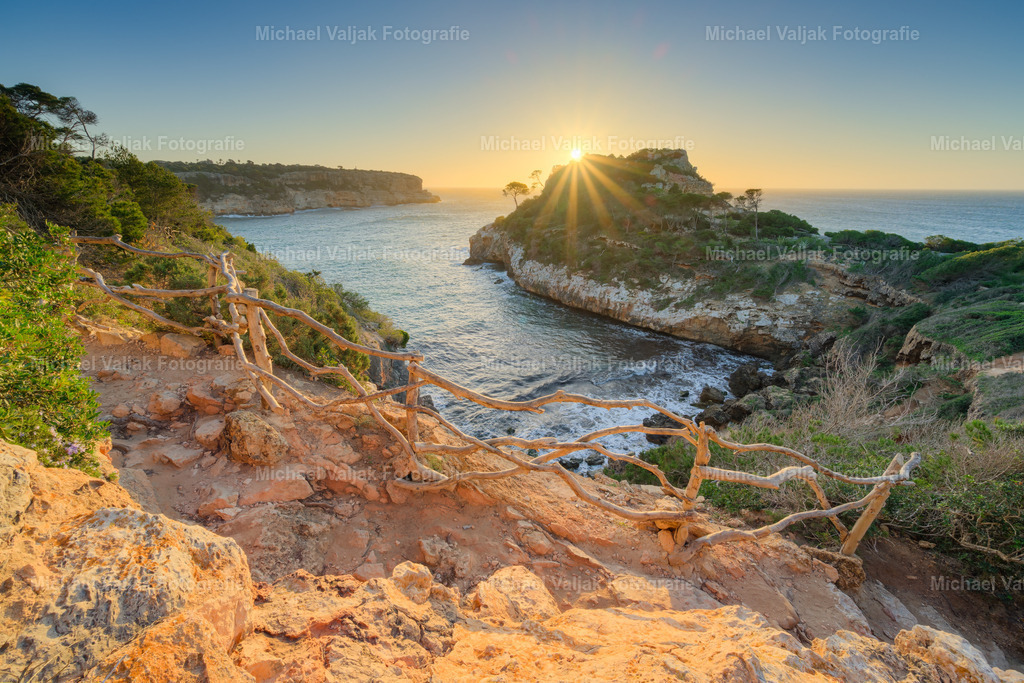 Caló des Moro auf Mallorca | Über der Caló des Moro steigt die Sonne langsam über die Felswände und taucht die enge Bucht in ein weiches, warmes Morgenlicht. Die steilen Klippen beginnen zu leuchten, während das Wasser darunter ruhig glitzert und erste Reflexe auf die Felsen wirft. Die geschützte Lage der Bucht verstärkt die Stille dieses Moments, in dem Licht, Farbe und Küstenform einen klaren, natürlichen Start in den Tag formen. - Realisiert mit Pictrs.com