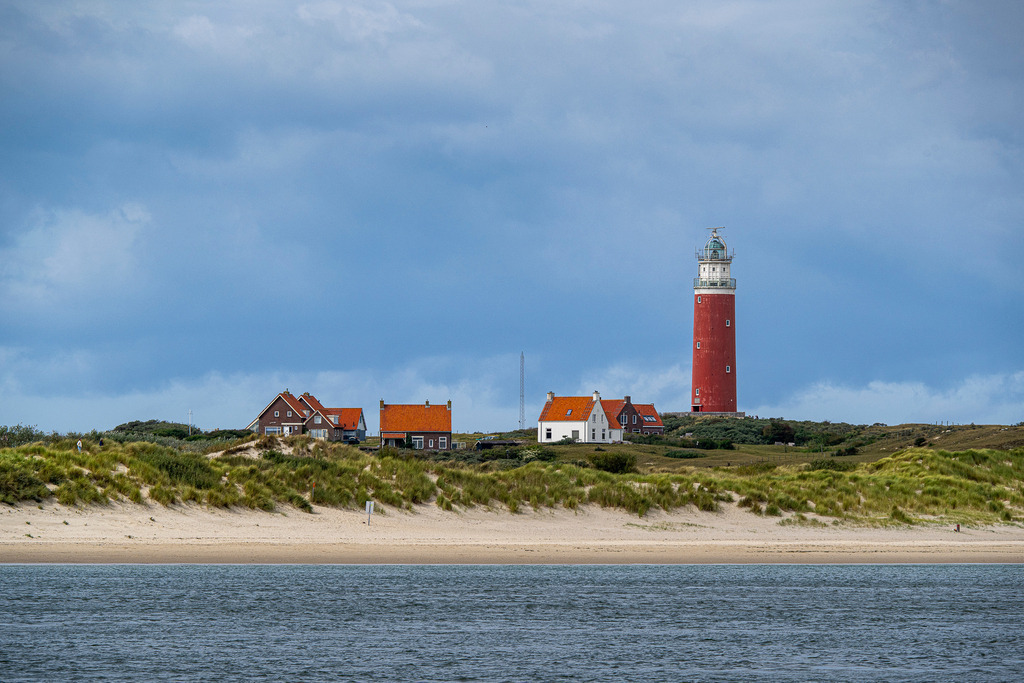 Leuchtturm Eierland auf Texel: Wahrzeichen an der Nordseeküste | Der roter Leuchtturm Eierland ragt im Norden der holländischen Westfriesischen Insel Texel hinter den Dünen empor. Im Vordergrund liegt der ruhige Strandstreifen an der Nordsee, während typische Küstenhäuser mit roten Ziegeldächern das Panorama ergänzen. Unter leicht bewölktem Himmel symbolisiert das maritime Motiv die idyllische Küstenlandschaft der Niederlande, ideal für Themen wie Tourismus, Nordseeurlaub, Reisekultur und Naturschutz. - Realisiert mit Pictrs.com