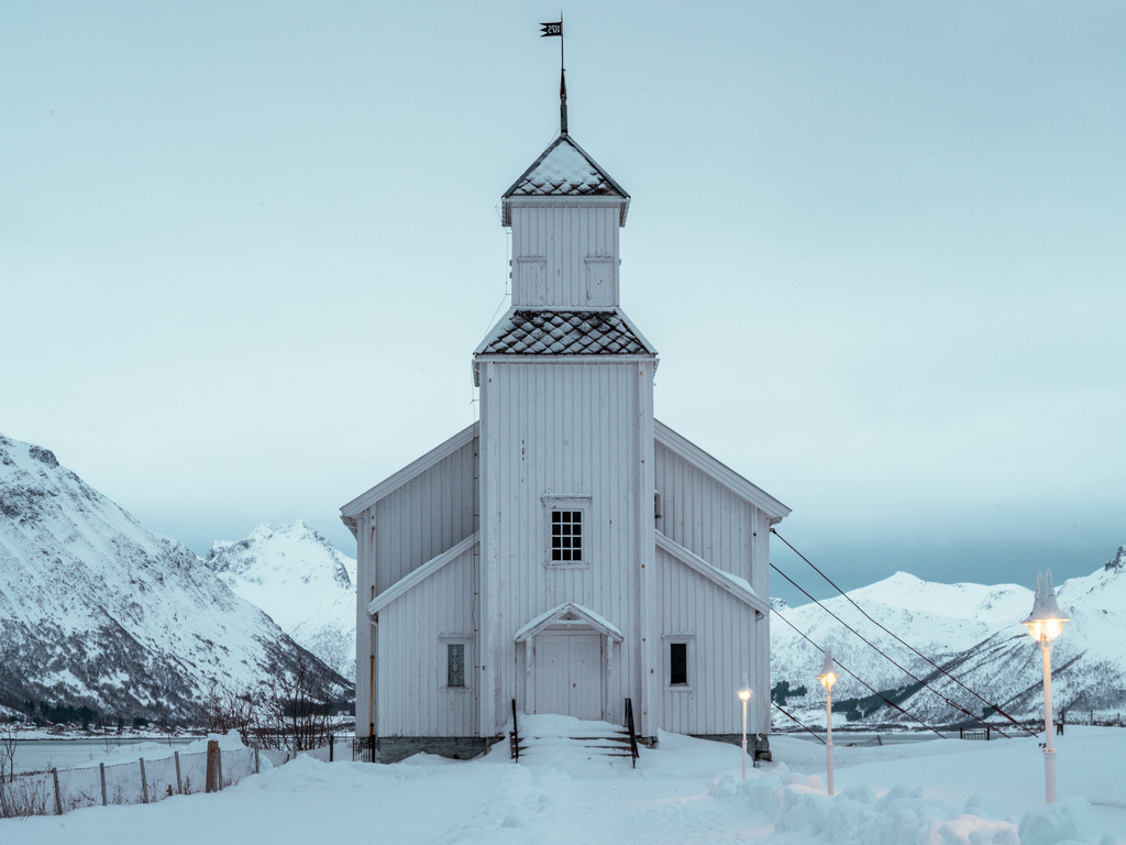 Norwegische Kirche in verschneiter Landschaft | Eine weiße Kirche steht ruhig in einer tief verschneiten Landschaft vor den Bergen Nordnorwegens. Die klare Architektur hebt sich dezent vom winterlichen Hintergrund ab. - Realisiert mit Pictrs.com
