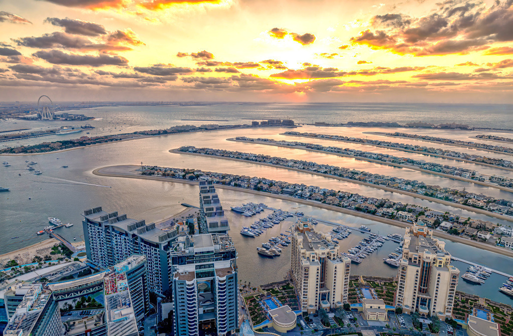 Die Palm Jumeirah in Dubai bei Sonnenuntergang | Das Bild zeigt einen Panoramablick auf die Palm Jumeirah in Dubai, aufgenommen von der Aussichtsplattform The View at The Palm. Diese Plattform befindet sich auf der 52. Etage des Palm Towers in einer Höhe von 240 Metern und bietet einen 360-Grad-Blick auf die künstliche Inselgruppe, den Arabischen Golf und die Skyline von Dubai. Highlights und Details zum StandortThe View at The Palm: Die Aussichtsplattform ist über das Einkaufszentrum Nakheel Mall direkt zugänglich, das mit dem Palm Tower verbunden ist.Sehenswürdigkeiten im Bild:Im Hintergrund links ist das Riesenrad Ain Dubai auf Bluewaters Island zu erkennen.Die Palmenwedel (Fronds) beherbergen zahlreiche Luxusvillen und Wohnanlagen. - Realisiert mit Pictrs.com