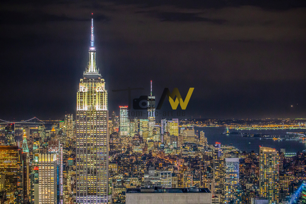 Skyline von New York-------Empire State Building bei Nacht | Das Bild zeigt eine Nachtaufnahme der Skyline von New York City mit dem beleuchteten Empire State Building im Vordergrund.Die Aufnahme zeigt das ikonische Empire State Building bei Nacht.Es bietet einen Panoramablick auf die beleuchtete Skyline von Manhattan und das Wasser im Hintergrund.Das Bild wurde wahrscheinlich von einer erhöhten Position wie dem Rockefeller Center (Top of the Rock) aufgenommen.Am Himmel sind einige Sterne und Wolken zu sehen. - Realisiert mit Pictrs.com