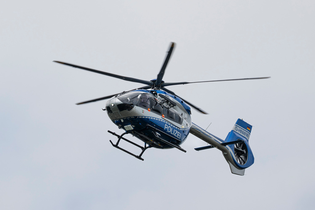 Polizeihubschrauber NRW | Das Foto präsentiert einen Airbus H145 (BK117 D-3) der nordrhein-westfälischen Polizei (Fliegerstaffel „Hummel“). Der Hubschrauber ist in der charakteristischen blau-silbernen Lackierung gehalten und trägt den deutlich sichtbaren Schriftzug „POLIZEI“. Das Luftfahrzeug ist mit einem Fünfblattrotor und einem geschlossenen Heckrotor (Fenestron) ausgestattet. Am Heck ist die Kennung D-HNWW sowie die Deutschlandflagge zu erkennen. Die Perspektive von schräg unten verleiht der Maschine eine dynamische und präsente Wirkung. - Realisiert mit Pictrs.com