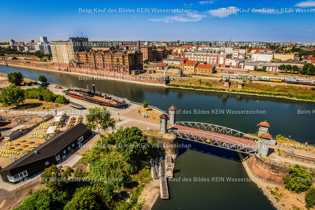 kleine Hubbrücke im Wissenschaftshafen | Das Foto zeigt die kleine Hubbrücke im Wissenschaftshafen Magdeburg aus einer bodennahen Perspektive. Die kompakte Stahlfachwerkkonstruktion mit ihrem markanten Hubportal vermittelt eindrucksvoll die Ingenieurtechnik historischer Hafenanlagen. Durch die niedrige Kameraposition treten die genieteten Stahlträger, der bewegliche Brückenüberbau und die technischen Details besonders deutlich hervor.Die Hubbrücke wurde Anfang des 20. Jahrhunderts als Teil der Hafeninfrastruktur errichtet und diente dem Anschluss der Gleisanlagen im Handelshafen. Über den elektrischen Hubmechanismus konnte der Brückenüberbau angehoben werden, sodass Schiffe den Hafenarm ungehindert passieren konnten. Hubbrücken dieser Bauart galten damals als besonders platzsparende Lösung, da sie im Gegensatz zu Dreh- oder Klappbrücken nur wenig Raum für ihre Bewegung benötigten. Die genietete Stahlkonstruktion ist ein typisches Beispiel des deutschen Stahlbrückenbaus jener Zeit.Die Aufnahme dokumentiert ein selten erhaltenes technisches Denkmal der Magdeburger Hafen- und Eisenbahngeschichte. Die kleine Hubbrücke erinnert an die enge Verbindung von Schifffahrt und Schienenverkehr und steht bis heute als Zeugnis der leistungsfähigen Hafenlogistik, die Magdeburg über viele Jahrzehnte zu einem bedeutenden Binnenhafen machte. - Realisiert mit Pictrs.com