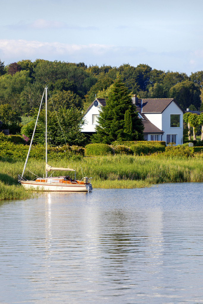 Wandbild: Segelboot am Schleiufer in Lindaunis | Dieses Wandbild im Hochformat zeigt ein Segelboot am Ufer der Schlei in Lindaunis. Auf der Schlei ist eine schöne Spiegelung des Segelboots zu sehen. Im Hintergrund befindet sich ein Haus am Waldrand.  - Realisiert mit Pictrs.com