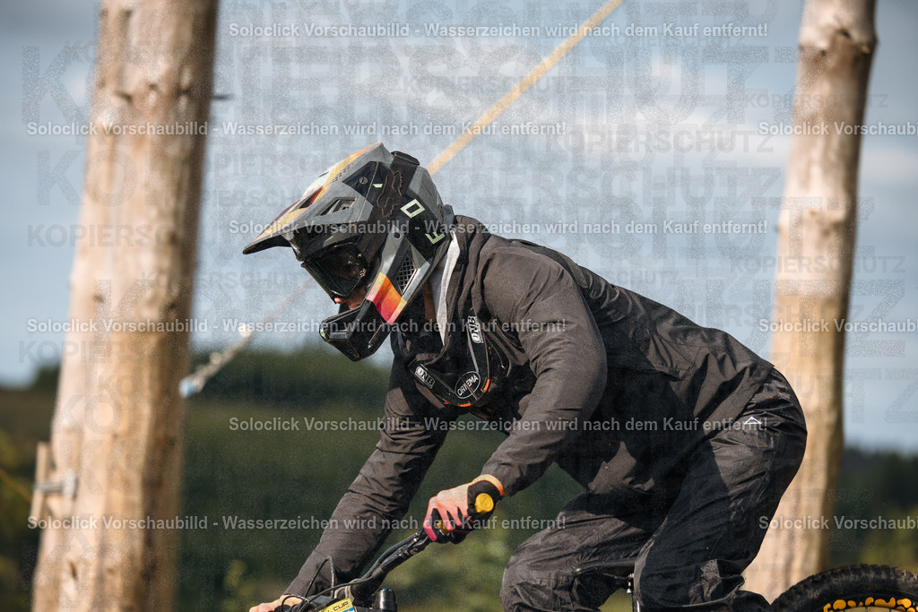 Mountainbiker in Aktion | Ein Mountainbiker in voller Montur fährt auf einem Trail. Er trägt einen schwarzen Helm mit Visier, eine schwarze Weste, schwarze Handschuhe und schwarze Hosen. Er fährt ein gelbes Mountainbike mit schwarzen Reifen. Der Hintergrund besteht aus Bäumen und Büschen. - Realisiert mit Pictrs.com
