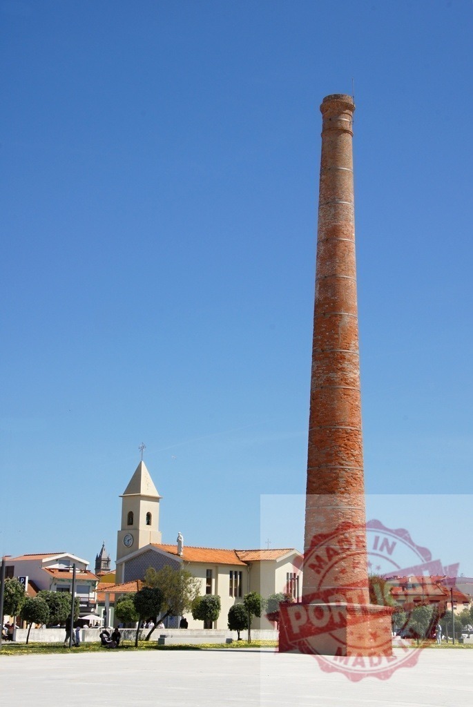 Old brick chimney standing tall in village square | Brick chimney and church tower standing in a sunny village square - Realized with Pictrs.com