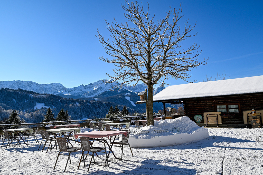 Wintermärchen auf der Höhe: Stille Einkehr auf der Pfeifferalm | Ein Anblick voller winterlicher Ruhe, Nostalgie und alpiner Geborgenheit, der das Herz wärmt: Frisch verschneit präsentiert sich die idyllische Terrasse der Pfeifferalm unter einem strahlend blauen, wolkenlosen Himmel. Die Tische und Stühle warten geduldig im Schnee auf die nächsten Sonnenanbeter, während ein knorriger, fast kahler Baum stolz im Zentrum aufragt. Daneben schmiegt sich die schneebedeckte Holzhütte harmonisch in die Landschaft. Im Hintergrund breitet sich ein atemberaubendes Panorama aus tief verschneiten Wäldern und den majestätischen, weißen Gipfeln des Wettersteingebirges aus. Diese Aufnahme fängt den Zauber eines klaren Wintermorgens in den Bergen perfekt ein – ein Moment der puren Entschleunigung und winterlichen Idylle. - Realisiert mit Pictrs.com