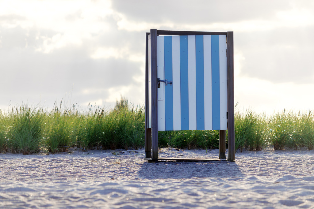 Leinwand: Umkleidekabine am Sandstrand | Dieses Wandbild im Querformat zeigt eine charmante Umkleidekabine am Strand von Weidefeld. Die Kabine, mit ihren blauen und weißen Streifen, steht malerisch auf dem Sand, während der Strandhafer im Hintergrund in der Unschärfe erscheint. Der bewölkte, aber sonnendurchflutete Himmel verleiht der Szene eine beruhigende und zugleich lebendige Atmosphäre. Dieses Wandbild eignet sich ideal, um jedem Raum eine maritime und erfrischende Note zu verleihen. - Realisiert mit Pictrs.com