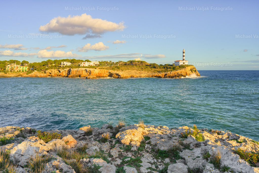 Blick zum Leuchtturm von Portocolom auf Mallorca | Der Blick richtet sich über die Küste von Portocolom hinweg zum Leuchtturm, der ruhig und beständig über dem Meer steht. Das mediterrane Licht betont die klaren Linien des Turms und lässt das Wasser in intensiven Blau‑ und Türkistönen leuchten.Die Szene wirkt gleichzeitig lebendig und gelassen – ein Moment, der die besondere Atmosphäre der mallorquinischen Ostküste einfängt und die Verbindung von Natur, Meer und maritimer Architektur spürbar macht. - Realisiert mit Pictrs.com