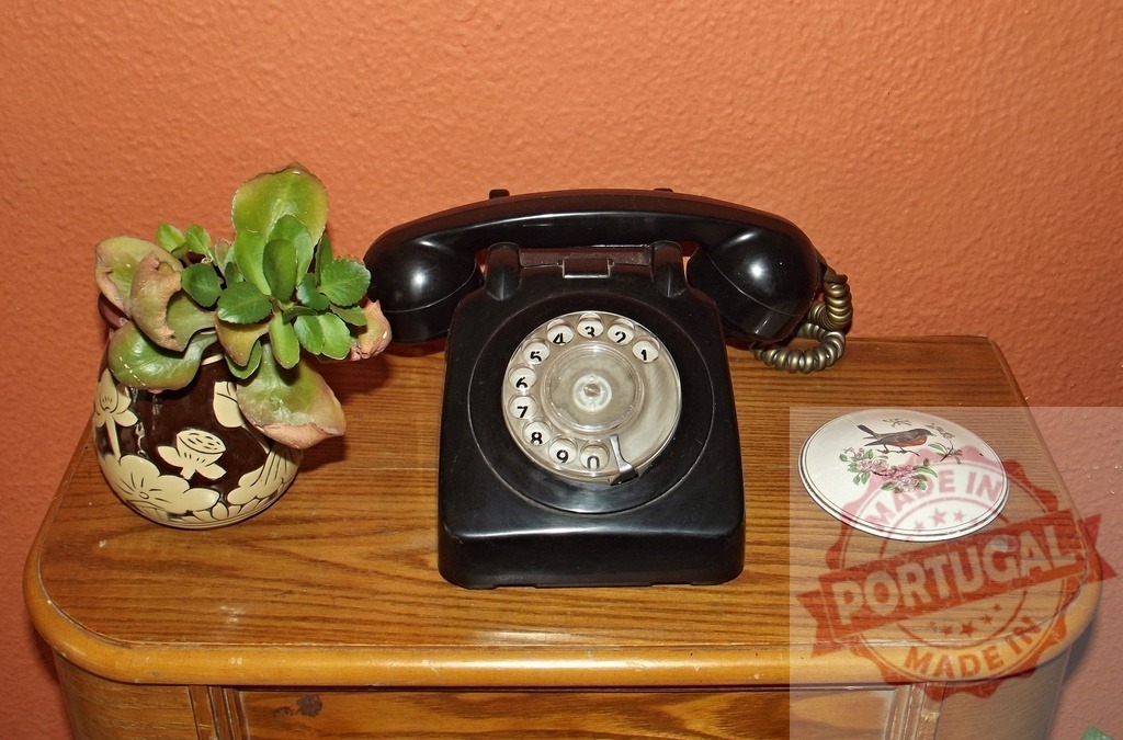 Old black rotary phone standing on wooden furniture with plant and trinket box | Vintage black rotary phone sits on wooden furniture next to a potted plant and a small trinket box, against an orange wall - Realized with Pictrs.com