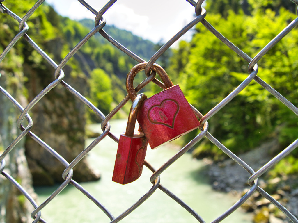 Schloss mit Herz | Zwei gravierte Vorhängeschlösser  hängen an einem Maschendrahtzaun. Im Hintergrund ein wunderschönes Flusstal - Realisiert mit Pictrs.com