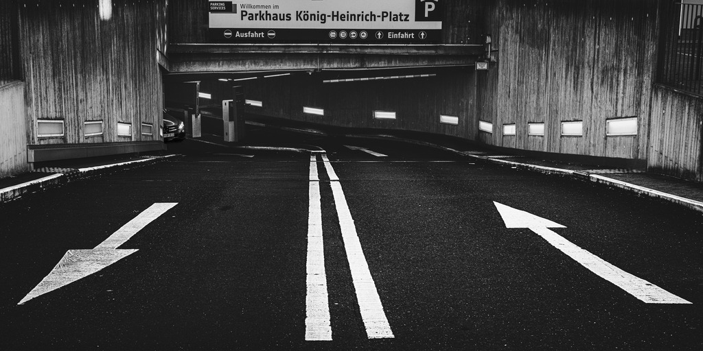 In or Out? | Düstere Schwarzweißaufnahme einer Tiefgarageneinfahrt am König-Heinrich-Platz in Duisburg. Die starken Kontraste, die glänzende Fahrbahn und die gegensätzlichen Pfeilrichtungen erzeugen eine spannungsgeladene urbane Atmosphäre. Das Bild spielt mit der Symbolik von Entscheidung, Bewegung und Richtung – hinein oder hinaus.Ein minimalistisches Fine-Art-Motiv mit cineastischer Ruhrpott-Ästhetik und klarer grafischer Wirkung. - Realisiert mit Pictrs.com