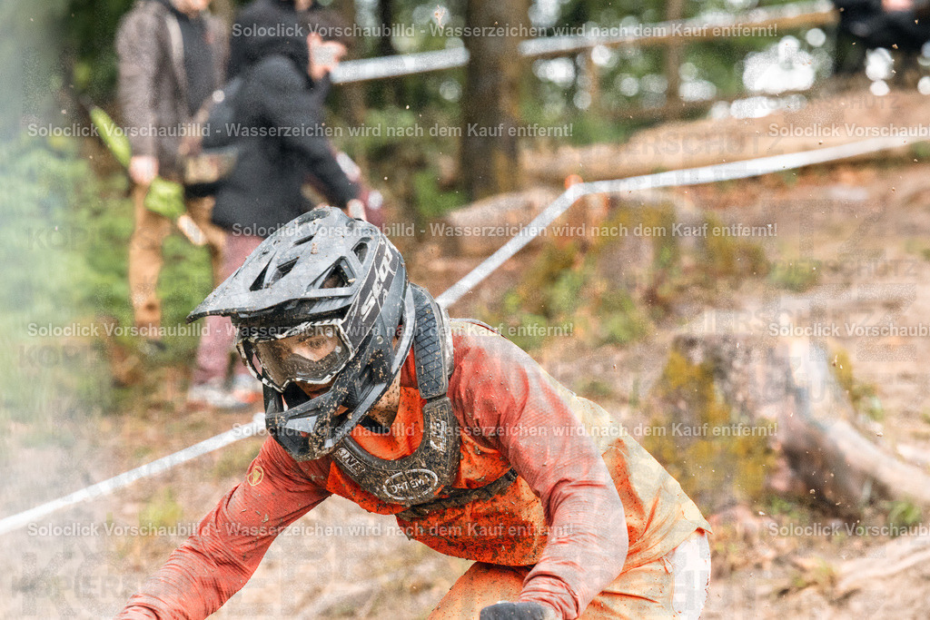 Mountainbike-Abenteuer im Gelände | Ein Mountainbiker in voller Ausrüstung, einschließlich Helm und Schutzbrille, kämpft sich einen steilen, schlammigen Hang hinab. Er trägt ein orangefarbenes Trikot und Handschuhe. Der Hintergrund besteht aus Bäumen und Vegetation. Der Fokus liegt auf dem Fahrer, der sich mit aller Kraft abmüht, den Hang zu bewältigen. - Realisiert mit Pictrs.com