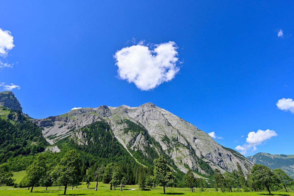 Ein Gruß der Natur: Wolkenherz über den Alpengipfeln | Ein Anblick, der das Herz erwärmt und ein tiefes Gefühl von Glück und Geborgenheit schenkt: Wie ein liebevoller Gruß des Himmels schwebt eine weiße Wolke in markanter Herzform direkt über dem mächtigen Felsmassiv des Karwendelgebirges. Die schroffen, grauen Kalksteinwände ragen majestätisch empor, sanft kontrastiert von saftig grünen Bergwäldern und den idyllischen Almwiesen des Großen Ahornbodens im Vordergrund. Vor dem makellosen, strahlenden Blau des Sommerhimmels wird diese Aufnahme zu einem poetischen Symbol für die Liebe zur Bergwelt. Ein magischer Moment voller Leichtigkeit, Romantik und zeitloser Schönheit, der zum Träumen einlädt. - Realisiert mit Pictrs.com