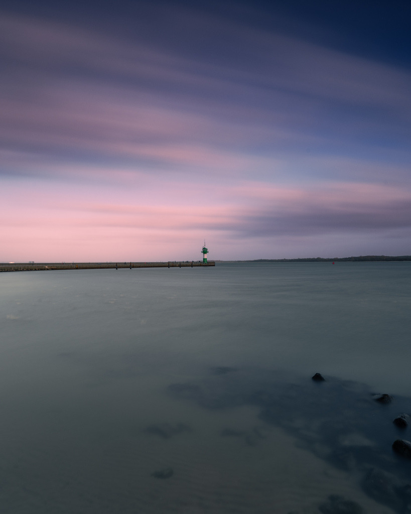 Molenfeuer im Abendlicht | Das grüne Molenfeuer steht am Horizont unter einem sanft gefärbten Abendhimmel. Durch die Langzeitbelichtung wirken Wasser und Wolken besonders ruhig und verleihen der Küstenszene eine minimalistische Atmosphäre. - Realisiert mit Pictrs.com