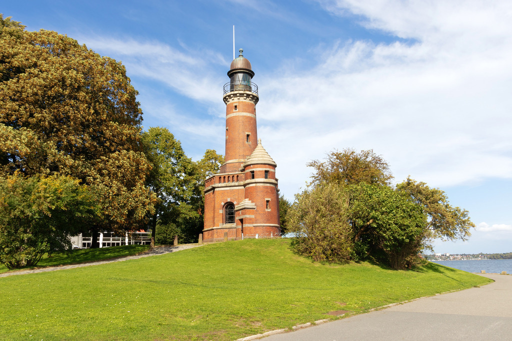 Wandbild: Leuchtturm in Kiel Holtenau | Das Wandbild im Querformat zeigt den Leuchtturm in Kiel Holtenau im Sommer. Der Leuchtturm an der Einfahrt zum Nord-Ostsee-Kanal befindet sich auf einem kleinen Hügel. Neben dem Leuchtturm stehen einige Bäume. Am blauen Himmel sind einige Wolken zu sehen.  - Realisiert mit Pictrs.com