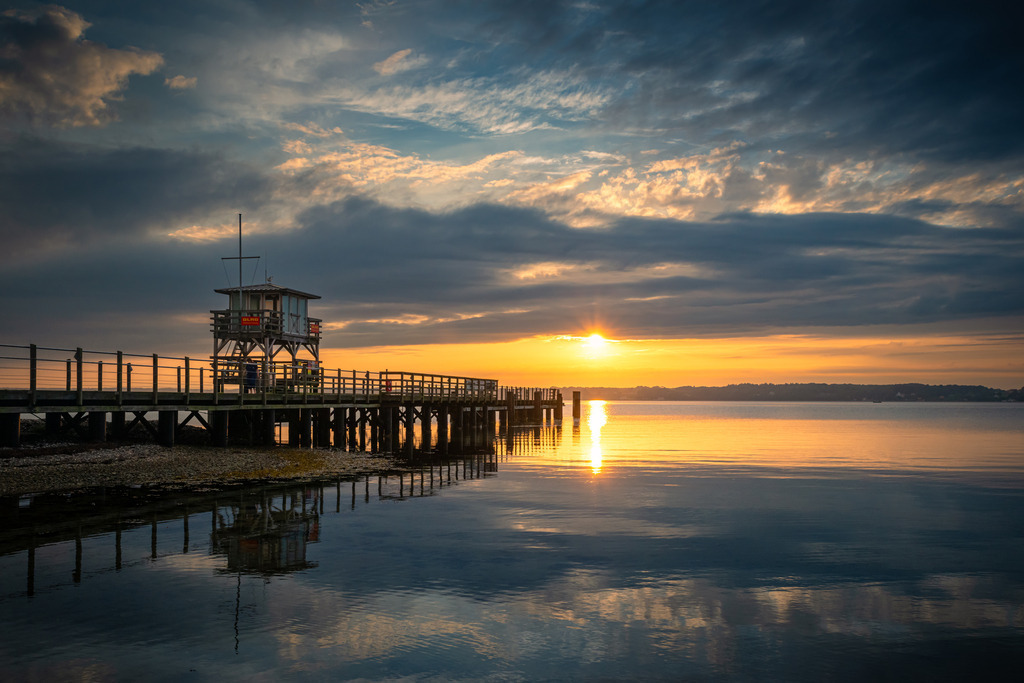 Seebrücke Glücksburg | Die Seebrücke in Glücksburg in der Abenddämmerung. Der Himmel spiegelt sich im ruhigen Wasser und verleihen dem Meer einen magischen Glanz.
Am Horizont verschmilzt der Himmel in einem Farbspektrum von Blau- und Orangetönen. Die Sonnenstrahlen spiegeln sich auf der Oberfläche des Wassers und verleihen dem Meer einen magischen Glanz.
Das Bild strahlt Ruhe und Gelassenheit aus und fängt den Moment der Stille und Schönheit ein. Es lädt den Betrachter ein, in die traumhafte Kulisse einzutauchen und die Schönheit dieses Ortes zu genießen.
