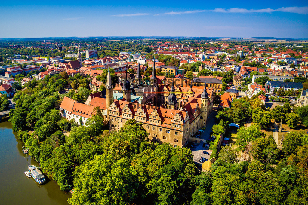 Schloss Merseburg in Sachsen-Anhalt an der Saale-0019 | Dieses Luftbild zeigt das Schloss Merseburg in Sachsen-Anhalt an einem sonnigen Sommertag aus einer eindrucksvollen Vogelperspektive. Die imposante Schlossanlage zählt zu den bedeutendsten Kulturdenkmalen Mitteldeutschlands und prägt gemeinsam mit dem Merseburger Dom seit Jahrhunderten die Silhouette der historischen Stadt an der Saale.Aus der Luft werden die beeindruckenden Dimensionen des Schlosses, die historischen Gebäudeflügel und die weitläufigen Schlossanlagen besonders deutlich sichtbar. Die Ursprünge der Anlage reichen bis in das frühe Mittelalter zurück. Über viele Jahrhunderte war Schloss Merseburg ein bedeutender Herrschafts- und Verwaltungssitz und gehört heute zu den wichtigsten Sehenswürdigkeiten Sachsen-Anhalts.Im Vordergrund der Aufnahme ist ein Abschnitt der Saale zu erkennen, die das Stadtbild von Merseburg seit Jahrhunderten prägt. Der Fluss bildet einen reizvollen Kontrast zur historischen Architektur und unterstreicht die besondere Lage der Stadt in der Saale-Auenlandschaft. Die sommerliche Vegetation, das warme Sonnenlicht und der klare Himmel verleihen der Szene eine freundliche und einladende Atmosphäre.Dieses Luftbild zeigt Schloss Merseburg als eines der bedeutendsten historischen Bauwerke Sachsen-Anhalts und vermittelt einen eindrucksvollen Eindruck von Geschichte, Architektur und Flusslandschaft. - Realisiert mit Pictrs.com