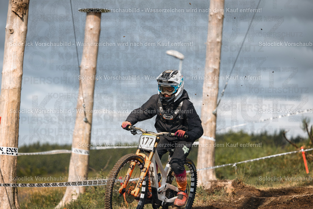 Mountainbike-Action | Ein Mountainbiker in voller Ausrüstung rast auf einem staubigen, unebenen Pfad hinunter. Er trägt einen schwarzen Helm mit weißen Akzenten, eine dunkle Brille und eine schwarze Jacke mit orangefarbenen Details. Seine Fahrerkleidung umfasst Handschuhe, Stiefel und eine Hose mit auffälligen Mustern. Das Mountainbike ist weiß mit orangefarbenen Akzenten und hat eine robuste Bauweise. Im Hintergrund sind Bäume und ein paar hölzerne Pfosten zu sehen, die den Kurs markieren. Ein Band markiert den Kurs. - Realisiert mit Pictrs.com