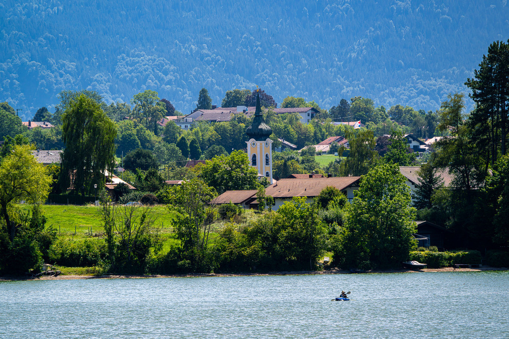 Malerische Kulisse in Seehausen: Kirche und Natur am Staffelsee | Ein traumhafter Panoramablick über das Blaue Land: Am Ufer des Staffelsees ragt der charakteristische Zwiebelturm der St.-Michael-Kirche in Seehausen pittoresk aus dem satten Grün hervor. Im Vordergrund zieht ein Kajakfahrer einsam seine Bahnen über das glitzernde Wasser, während im Hintergrund die bewaldeten Hänge der Voralpen emporragen. Diese friedliche Szenerie fängt die verträumte Magie Oberbayerns perfekt ein – ein Ort voller Harmonie, Tradition und gelebter Entschleunigung. - Realisiert mit Pictrs.com