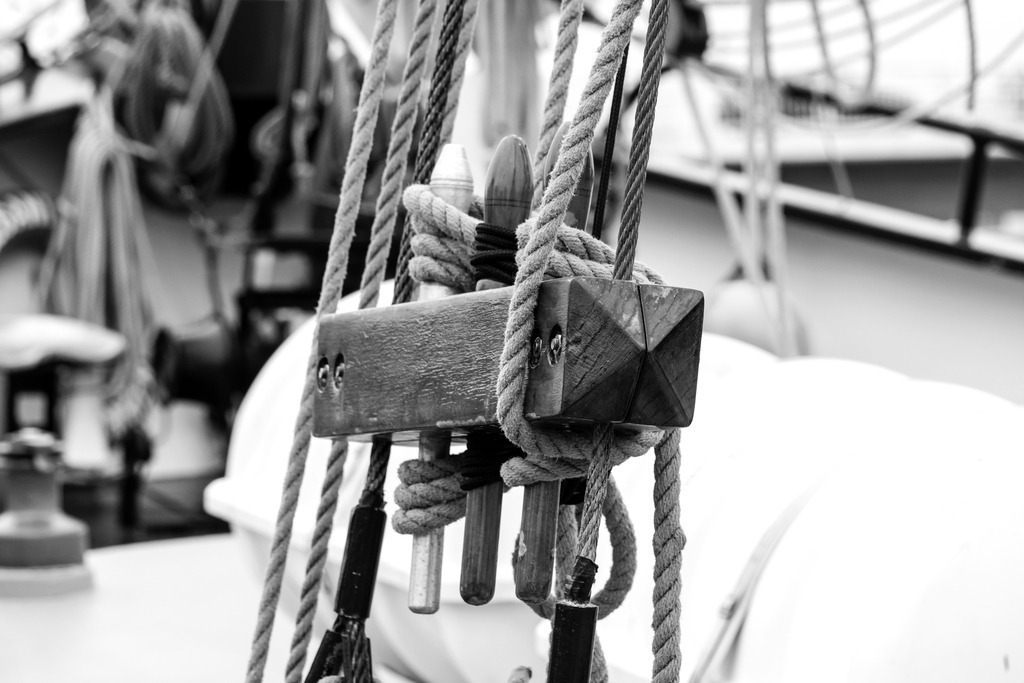 Wandbild: Detailansicht eines Segelschiffs in Kappeln an der Schlei | Erleben Sie die meisterhafte Handwerkskunst und die maritime Eleganz mit dieser Schwarz-Weiß-Aufnahme eines Segelschiffs. Die Nahaufnahme zeigt einen hölzernen Block, durch den mehrere Seile laufen, und betont die Präzision und Sorgfalt im Umgang mit der Takelage. Die Seile sind ordentlich gewickelt und verknotet und vermitteln ein Gefühl von Ruhe und Beständigkeit.Dieses Bild, erhältlich auf Leinwand, Alu-Dibond, Acrylglas, Fineart und als Akustikbild, bringt die faszinierende Welt der Seefahrt und die technischen Feinheiten direkt in Ihr Zuhause. - Realisiert mit Pictrs.com