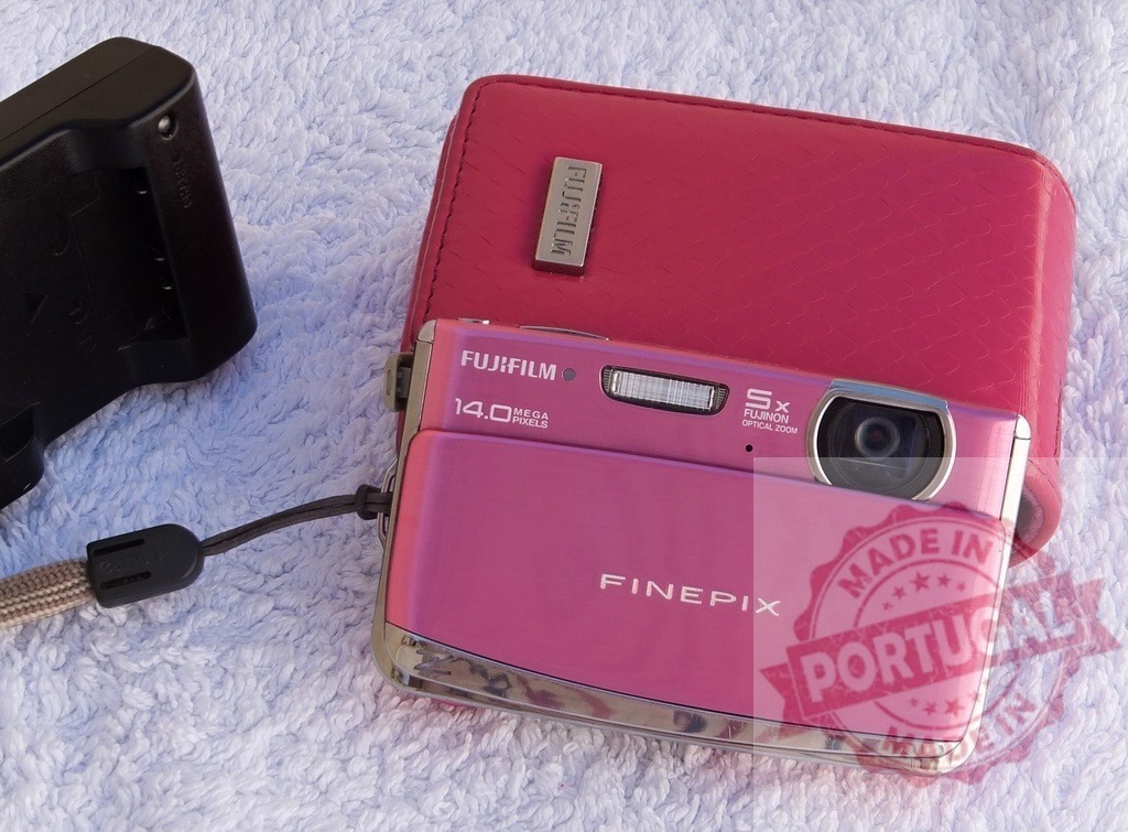 Pink Fujifilm FinePix camera with case and battery charger | Pink Fujifilm FinePix digital camera resting on a case with a battery charger nearby - Realized with Pictrs.com