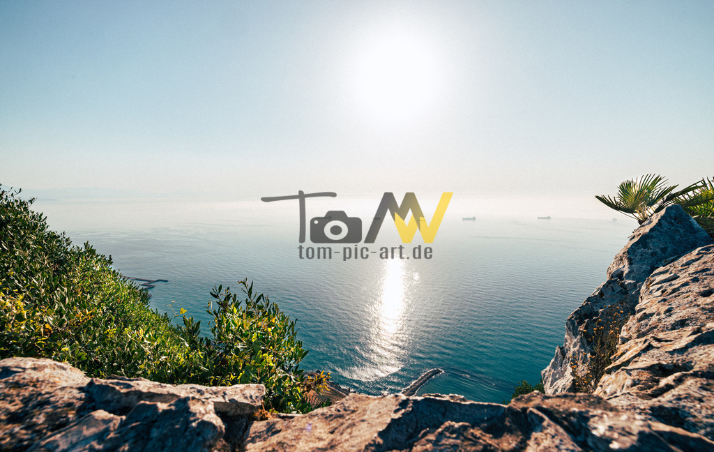 Blick auf die vorbei fahrenden Schiff vor Gibraltar | Das Bild zeigt eine Landschaftsaufnahme einer Küste oder Klippe mit Blick auf das Meer bei hellem Sonnenschein.Die Aufnahme wurde von einem erhöhten Standpunkt aus gemacht, wahrscheinlich einer Klippe oder einem Berg.Im Vordergrund sind Felsen und grüne Vegetation zu sehen. - Realisiert mit Pictrs.com