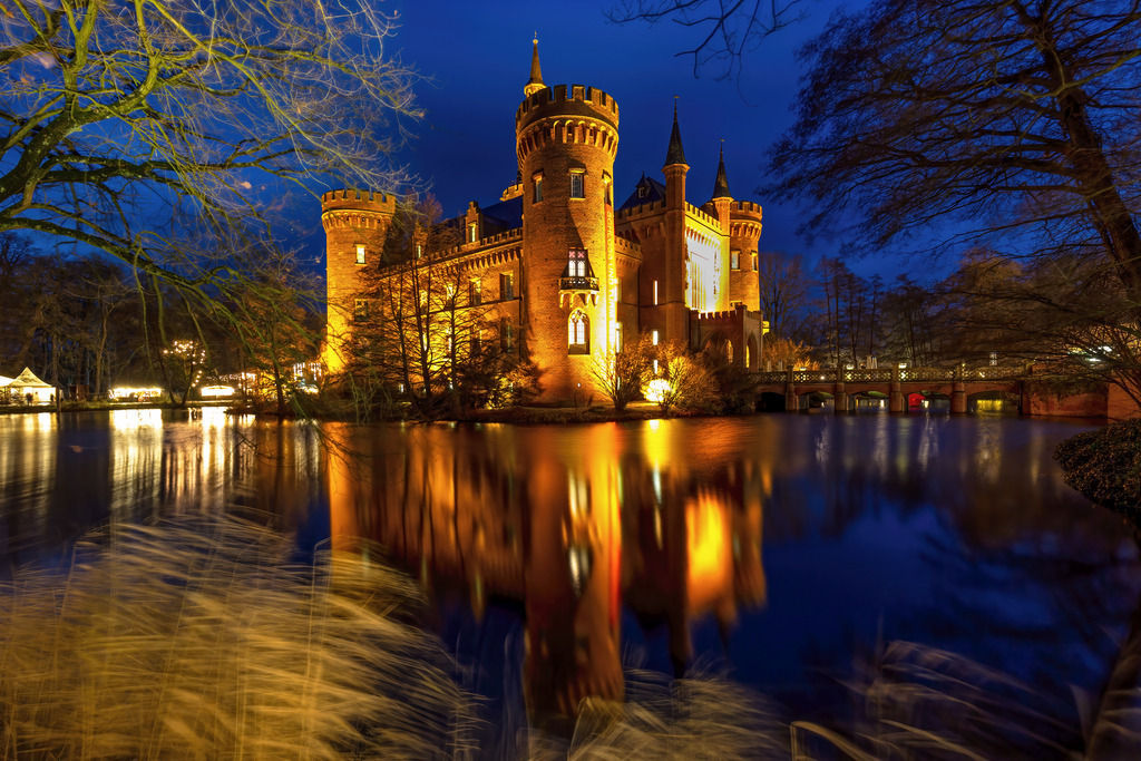 Wandbild - Märchenhaftes Schloss Moyland: Ein nächtliches Juwel | Dieses bezaubernde Bild zeigt Schloss Moyland bei Nacht, ein historisches Wasserschloss in Nordrhein-Westfalen, Deutschland. Die prachtvoll beleuchteten Türme und Mauern des Schlosses spiegeln sich im stillen Wasser des umgebenden Grabens wider, was eine märchenhafte Atmosphäre erzeugt. Die warmen Lichter des Schlosses kontrastieren wunderschön mit dem tiefblauen Nachthimmel und den schattigen Bäumen im Vordergrund. Der Schein der Lichter auf dem Wasser und die friedliche Umgebung verleihen diesem Bild eine romantische und zeitlose Qualität.