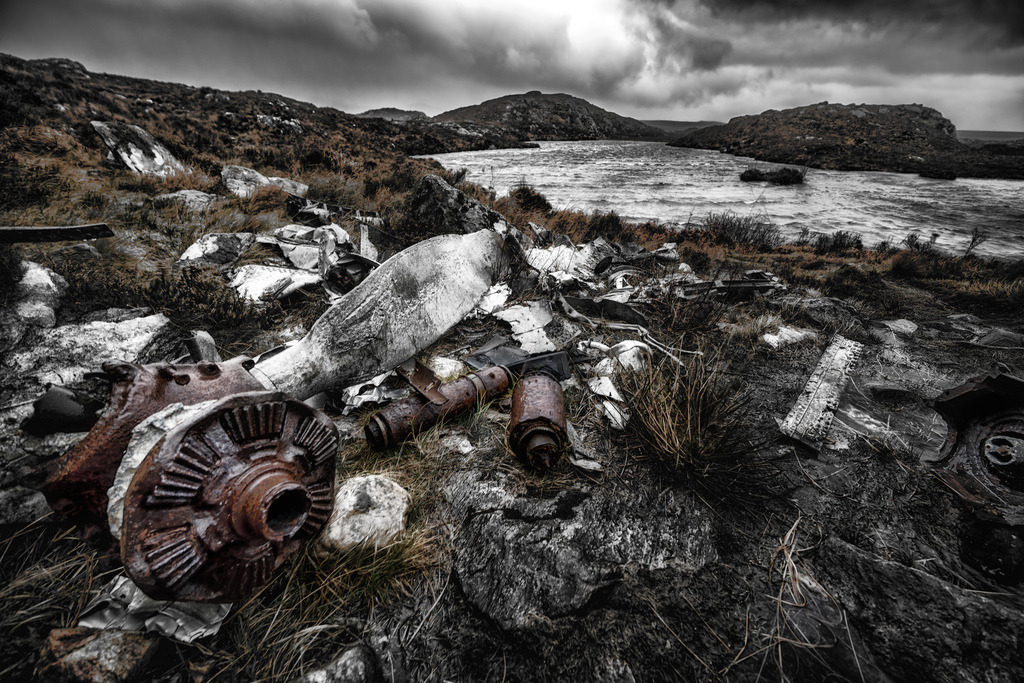 Absturzstelle eines USAAF B-24H Liberator-Bombers | Am 13. Juni 1945 stürzte ein USAAF B-24H Liberator nahe Gairloch, Schottland, ab – alle 15 Besatzungsmitglieder starben. Das Flugzeug war von Prestwick in die USA unterwegs, verliess den Kurs vermutlich wegen Navigationsfehler, Technik oder Wetter und stürzte beim Versuch einer Notlandung im Loch Gairloch in die Hügel oberhalb der Fairy Lochs ab, nachdem es den Berg Slioch streifte. Die Trümmer liegen heute verstreut am Ufer und auf dem Hügel; einige Teile wie Rumpf, Propeller und Triebwerke sind noch erkennbar. Der Ort ist eine Kriegsgräberstätte mit Gedenktafel und Besucher-Gedenkobjekten.Rostige Metallteile und zerbrochene Flugzeugtrummer liegen verstreut auf dem felsigen Ufer eines Sees in Gairloch, Schottland. Die dramatische Komposition mit einem weiten Blickwinkel betont die raue Landschaft und den dunklen, bewolkten Himmel. Die selektive Farbgebung hebt die verwitterten Wrackteile hervor und vermittelt eine melancholische Stimmung. - Realisiert mit Pictrs.com