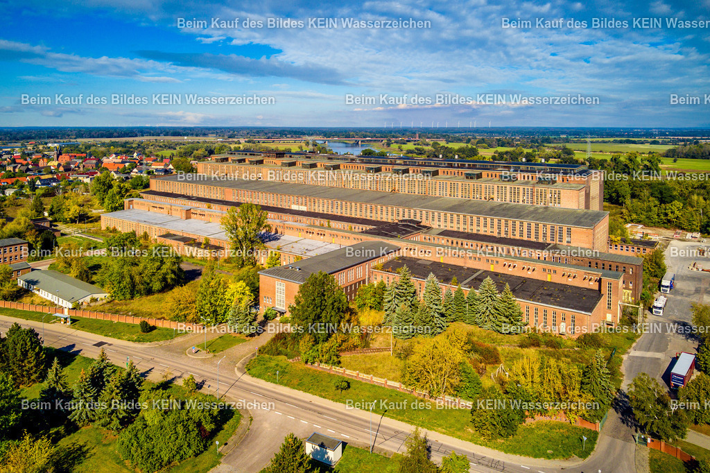 Kraftwerk Vockerode Elbe in Wittenberg-0014 | Das Luftbild zeigt das ehemalige Kraftwerk Vockerode aus einer Drohnenperspektive an einem sonnigen Sommertag. Der klare blaue Himmel hebt die markante Industriearchitektur eindrucksvoll hervor und lässt die langgestreckte Maschinenhalle sowie die Kesselhäuser besonders deutlich erkennen. Aus der Vogelperspektive werden die Dimensionen des historischen Kraftwerks und seine Lage unmittelbar an der Elbe sichtbar.Mit dem Bau des Braunkohlekraftwerks wurde 1937 begonnen. Die ersten sechs Kraftwerksblöcke verfügten über eine Leistung von jeweils 35 Megawatt, später wurde die Anlage um weitere zwölf Blöcke mit jeweils 32 Megawatt erweitert. Ab 1968 versorgte das Kraftwerk die Stadt Dessau zusätzlich über eine 15 Kilometer lange Fernwärmeleitung mit Wärme. Die markanten vier Schornsteine waren jeweils 140 Meter hoch und prägten jahrzehntelang das Landschaftsbild, bis sie 2001 gesprengt wurden. Die Drohnenaufnahme dokumentiert eines der bedeutendsten Industriedenkmäler Sachsen-Anhalts. Trotz der Stilllegung in den 1990er-Jahren vermittelt das erhaltene Kraftwerksgebäude bis heute einen eindrucksvollen Eindruck von den Dimensionen und der Leistungsfähigkeit der historischen Energieerzeugung im mitteldeutschen Braunkohlerevier. - Realisiert mit Pictrs.com
