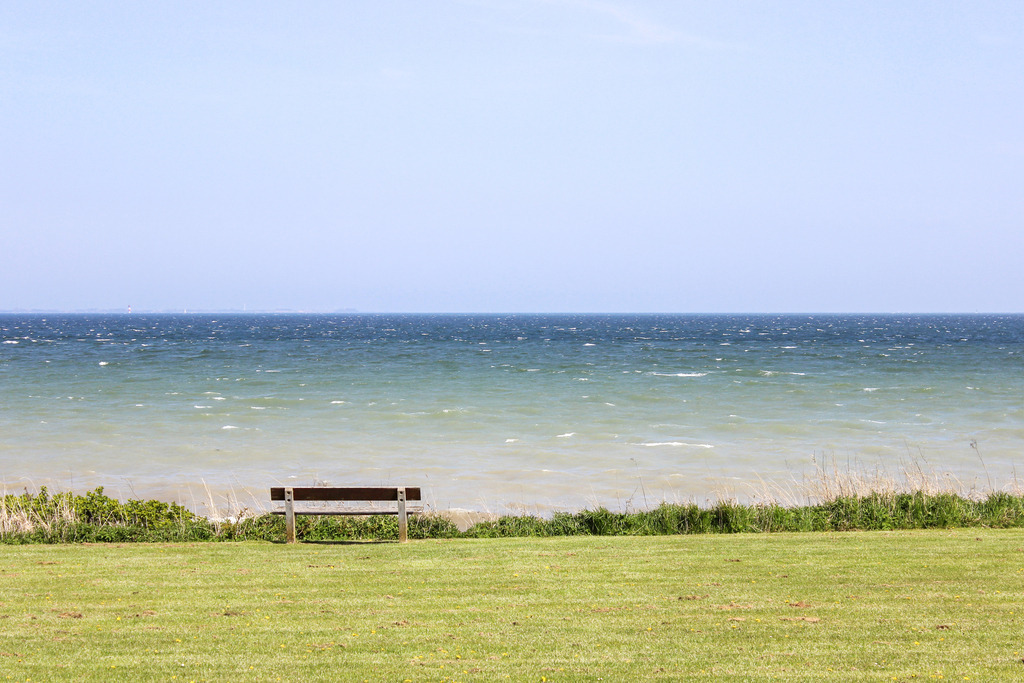Wandbild: Bank am Meer in Norgaardholz | Dieses Wandbild im Querformat zeigt die Küste in Norgaardholz. Direkt an der kleinen Steilküste am Strand befindet sich eine Bank zum verweilen. Auf dem Meer sind einige Wellen zu sehen und der blaue Himmel ist wolkenlos. - Realisiert mit Pictrs.com