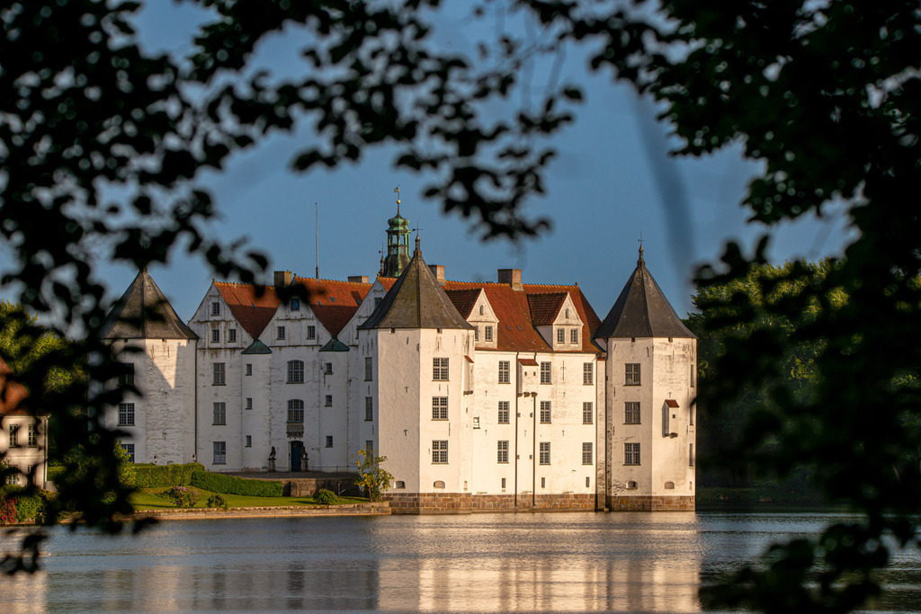 Schloss Glücksburg | Das Bild zeigt das majestätische Schloss in Glücksburg, welches Nachts mit den Farben von Schleswig-Holstein angestrahlt wird. Die warmen Farben der Beleuchtung lassen das Schloss in einem atemberaubenden Licht erstrahlen und betonen seine prächtigen Türme und Fassaden. Die sanfte Beleuchtung reflektiert sich im Schlossteich und schafft eine zauberhafte Atmosphäre, die den Betrachter in eine Welt voller Magie und Schönheit entführt. Die ruhige Wasseroberfläche lässt das Schloss wie in einem Spiegelbild erscheinen und erzeugt eine fast surreal wirkende Stimmung. Die Szenerie lädt zum Träumen und Verweilen ein. Es erinnert an die Geschichte und Bedeutung des Schlosses in Glücksburg als bedeutendes Kultur- und Geschichtsdenkmal der Region Schleswig-Holstein.
