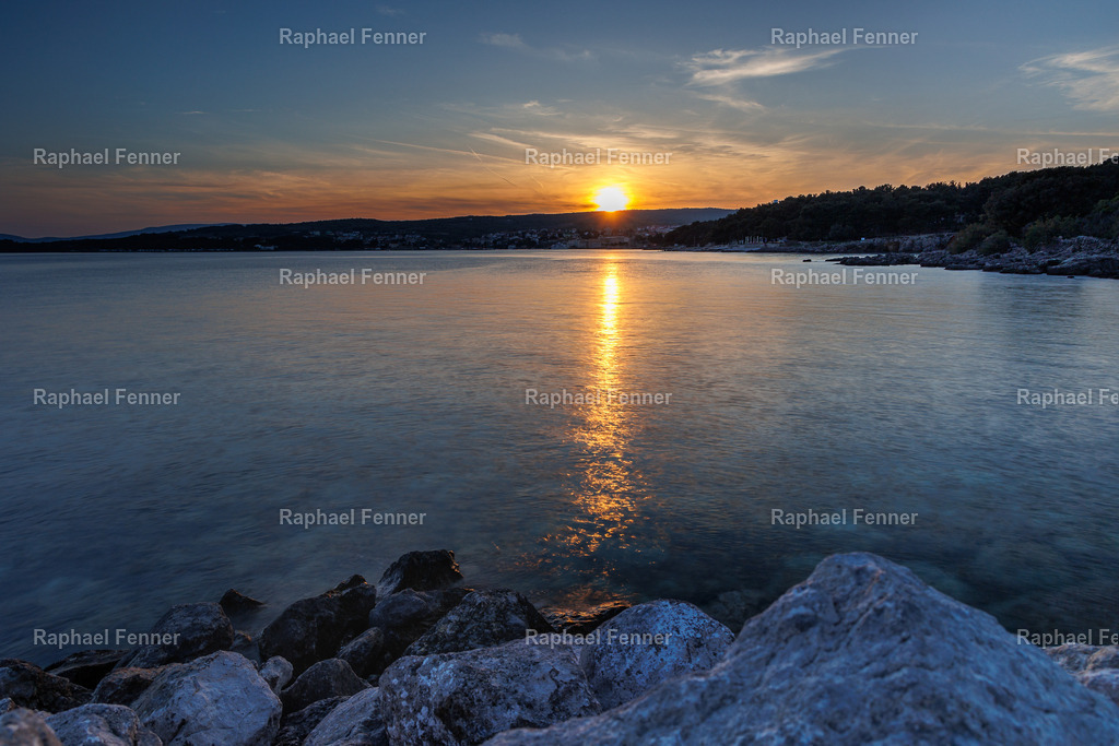 Sonnenuntergang auf Krk – Inselblick in Gold | Aufgenommen auf der Insel Krk mit Blick in Richtung der Stadt Krk. Die untergehende Sonne spiegelt sich golden auf dem ruhigen Meer, während die Felsen im Vordergrund die Szene erden. Eine friedliche Abendstimmung, die mediterrane Weite und Wärme ausstrahlt – ideal als Fine Art Print für dein Zuhause oder Büro.