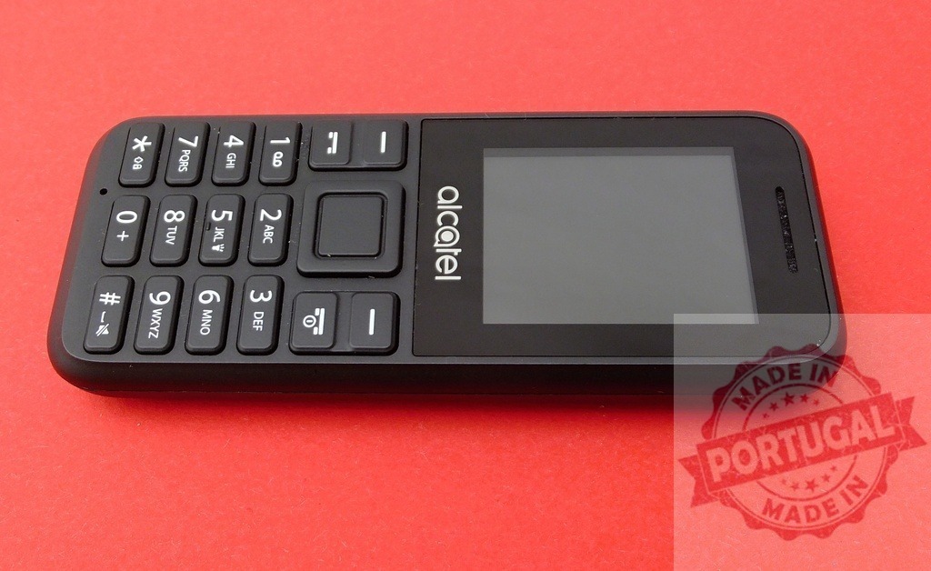 Alcatel mobile phone lying on red background | Black Alcatel basic mobile phone with buttons and blank screen lying on a red background - Realized with Pictrs.com
