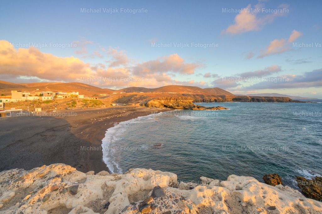 Playa de Ajuy auf Fuerteventura | Am Playa de Ajuy liegt die Abendsonne warm über der dunklen Lavaküste und taucht den Strand in ein weiches, goldenes Licht. Die Wellen rollen gleichmäßig an den schwarzen Sand, und das Meer schimmert in tiefen Blau‑ und Türkistönen, die im Kontrast zum dunklen Gestein noch intensiver wirken. Die Felsen entlang der Bucht leuchten in warmen Orangetönen, während die Sonne langsam tiefer sinkt und die Szene eine ruhige, fast zeitlose Stimmung bekommt. Ein Moment, in dem die raue Westküste Fuerteventuras ihre sanfte Seite zeigt. - Realisiert mit Pictrs.com