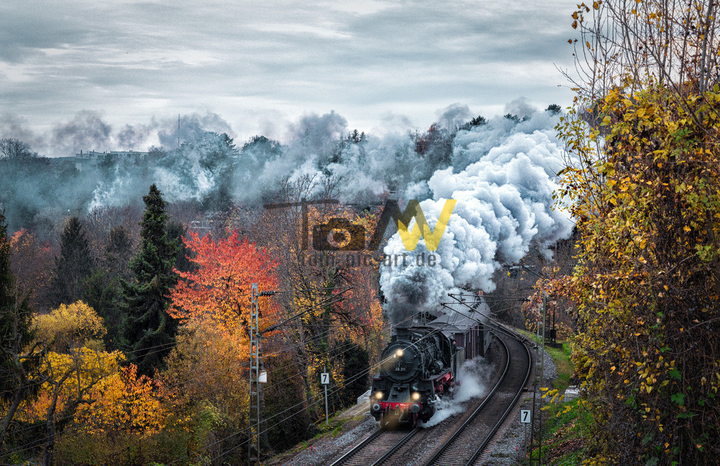 Dampflok fährt durch Stuttgart Münster auf Schusterstrecke | Das Bild zeigt eine Dampflokomotive, die durch eine malerische Herbstlandschaft fährt. Die Lokomotive stößt große Mengen Dampf und Rauch aus, was typisch für den Betrieb mit Kohle ist. Die Szene findet auf einer eingleisigen Bahnstrecke statt, umgeben von Bäumen m - Realisiert mit Pictrs.com